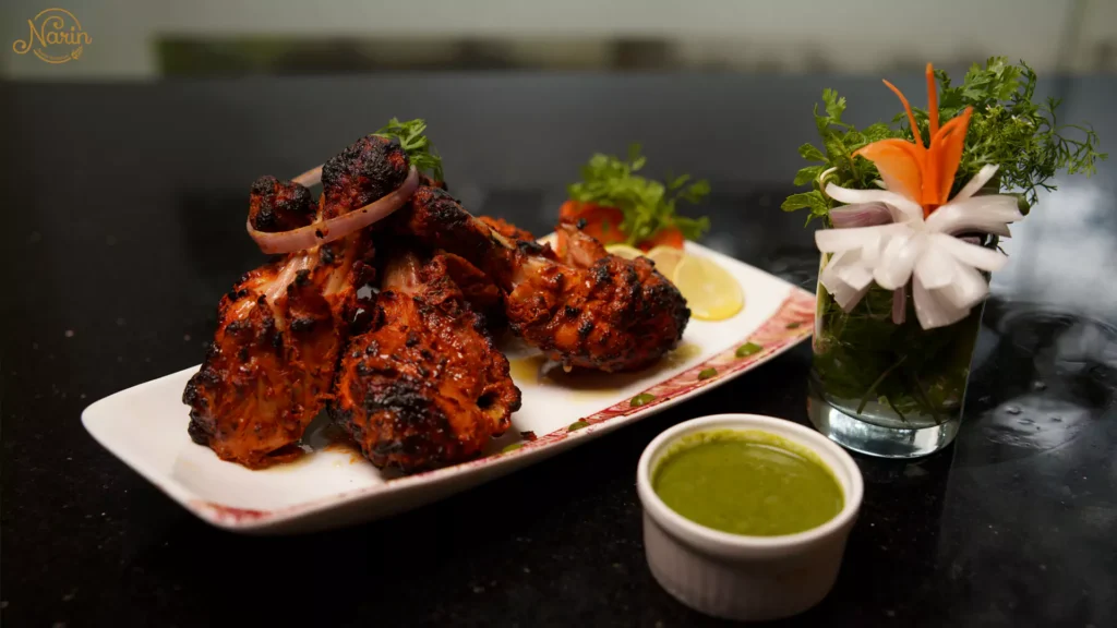 Narin Persian Restaurant presentation: marinated Persian-style grilled chicken drumsticks (Kebab-e Morgh) on a long plate, served with a fresh lime and coriander chutney, mint sprigs, and an intricate radish flower garnish, on a black marble table.