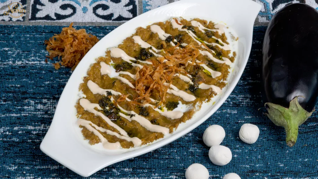 Top-down view of a white oval dish filled with Kashk Bademjan, a creamy Persian eggplant dip, garnished with swirls of white kashk, dark mint oil, and crispy golden fried onions. A raw eggplant and dried round kashk pieces sit nearby on a blue traditional fabric.
