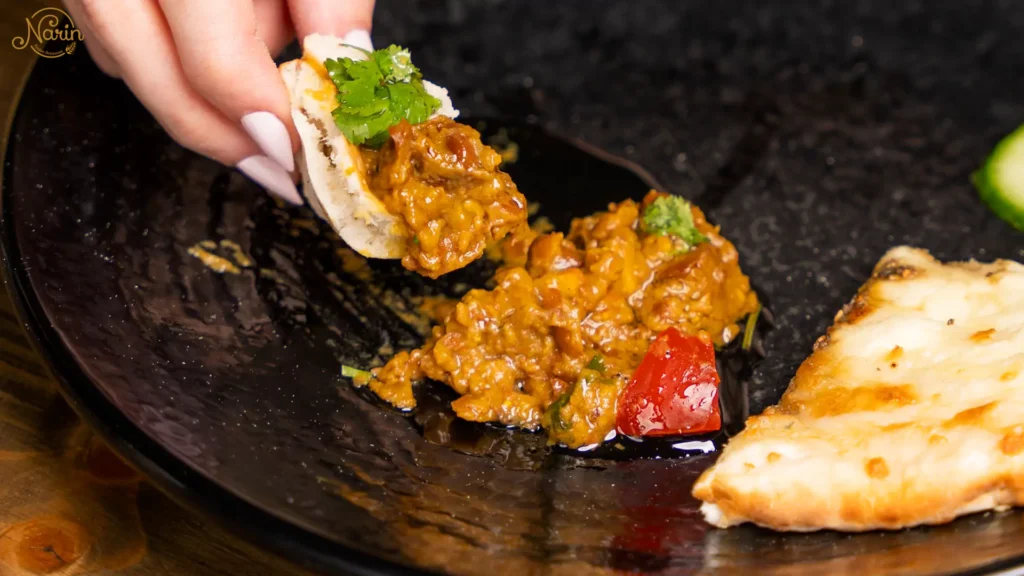 Close-up of a hand using a piece of traditional flatbread to scoop up Mirza Ghasemi, a vibrant smoky Persian appetizer made of fire-roasted eggplants, tomatoes, and garlic, served on a black plate with fresh herbs.