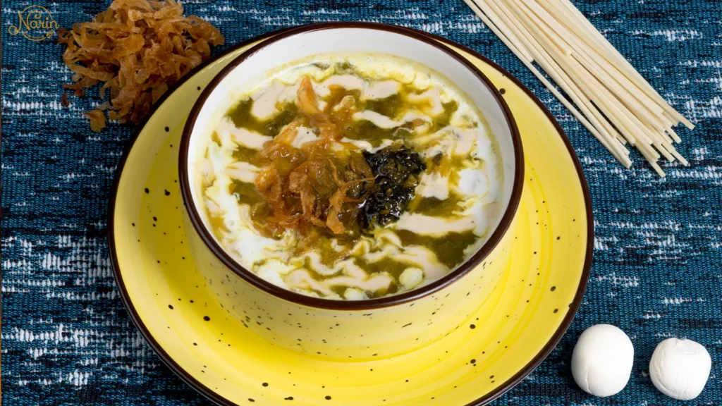 A yellow ceramic bowl filled with Ash Reshteh, a hearty Persian noodle and herb soup, topped with decorative swirls of white kashk, dark mint oil, and crispy fried onions. Uncooked wheat noodles and dried kashk pieces are placed next to the bowl on a blue patterned tablecloth.