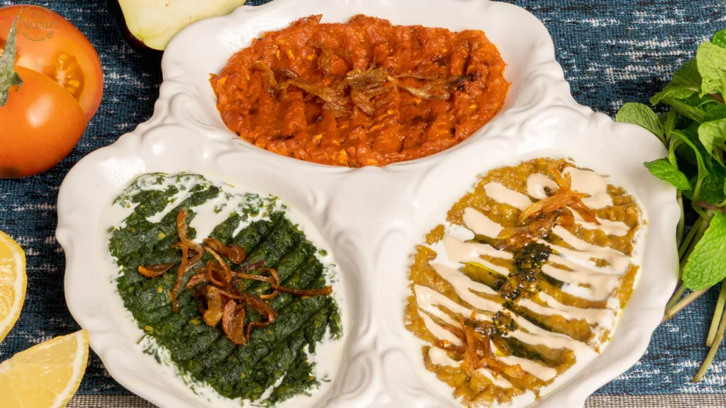 A three-compartment white serving dish showcasing a colorful spread of Persian appetizers, including smoky Mirza Ghasemi at the top, creamy Kashk Bademjan drizzled with kashk on the right, and a vibrant green spinach and yogurt dip on the left, all garnished with crispy fried onions and served alongside fresh mint and sliced tomatoes.