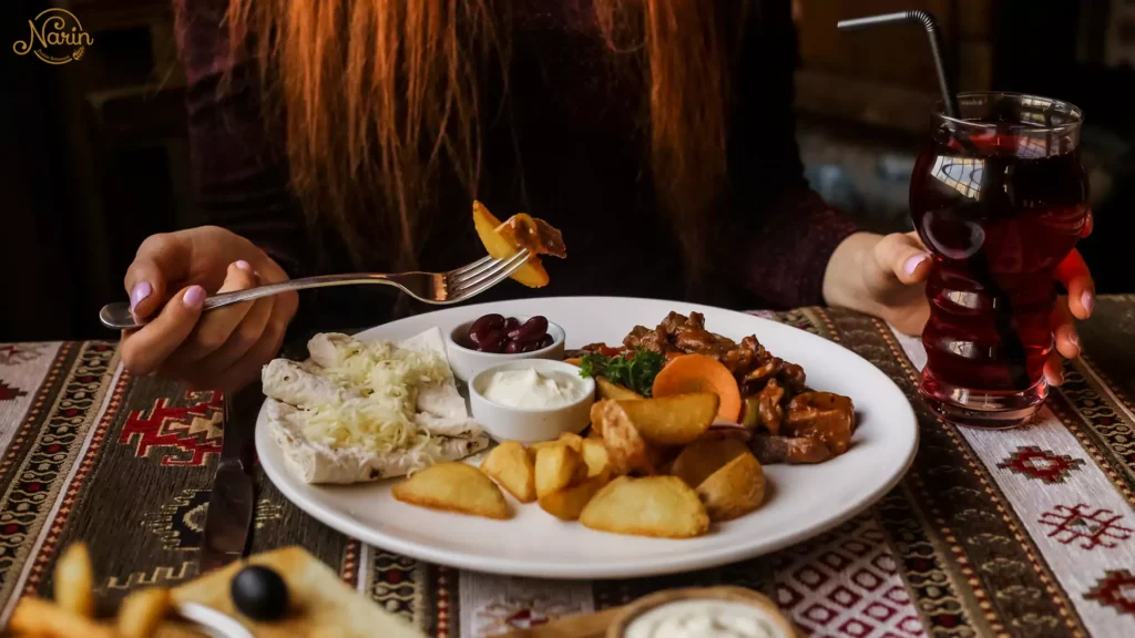 A close-up view of a woman with long red hair holding a fork with a bite of food. Her other hand holds a tall glass of dark red liquid with a straw. On the patterned traditional tablecloth is a plate with roasted potato wedges, meat chunks, two stuffed rolls with grated cheese, and small dishes of dip and olives. The edge of a bowl with more fries is in the foreground.