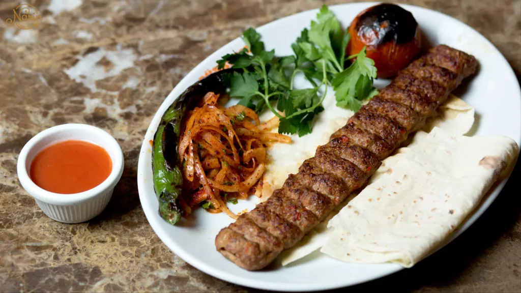Authentic Persian Koobideh kebab served over flatbread at Narin Persian Restaurant in Sydney, beautifully plated with a roasted tomato, charred green pepper, seasoned sliced onions, and fresh parsley, accompanied by a small bowl of red dipping sauce on a marble table.