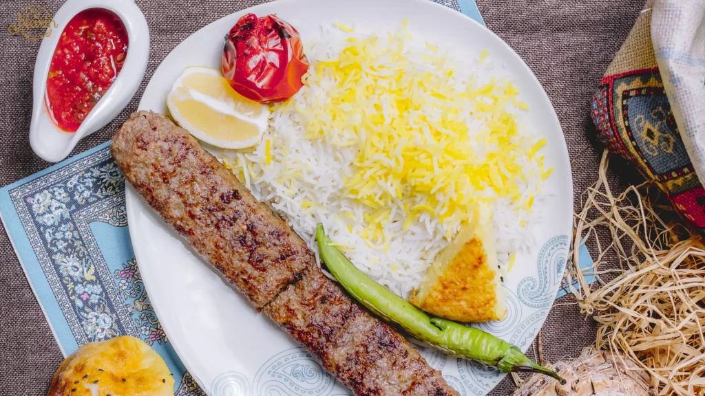 Top-down view of a traditional Persian Koobideh kebab plate featuring a grilled minced meat skewer alongside fluffy white and saffron rice, a charred tomato, a roasted green pepper, a lemon wedge, and a piece of crispy Tahdig, served with a side of red sauce.