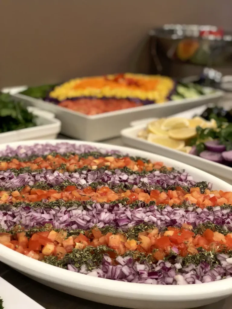 Close-up of a large catering platter at Narin Persian Restaurant in Sydney, featuring finely diced tomatoes and red onions beautifully arranged in alternating diagonal stripes and garnished with dried herbs. Other fresh salads and side dishes with lemon wedges are visible in the blurred background on the buffet table.