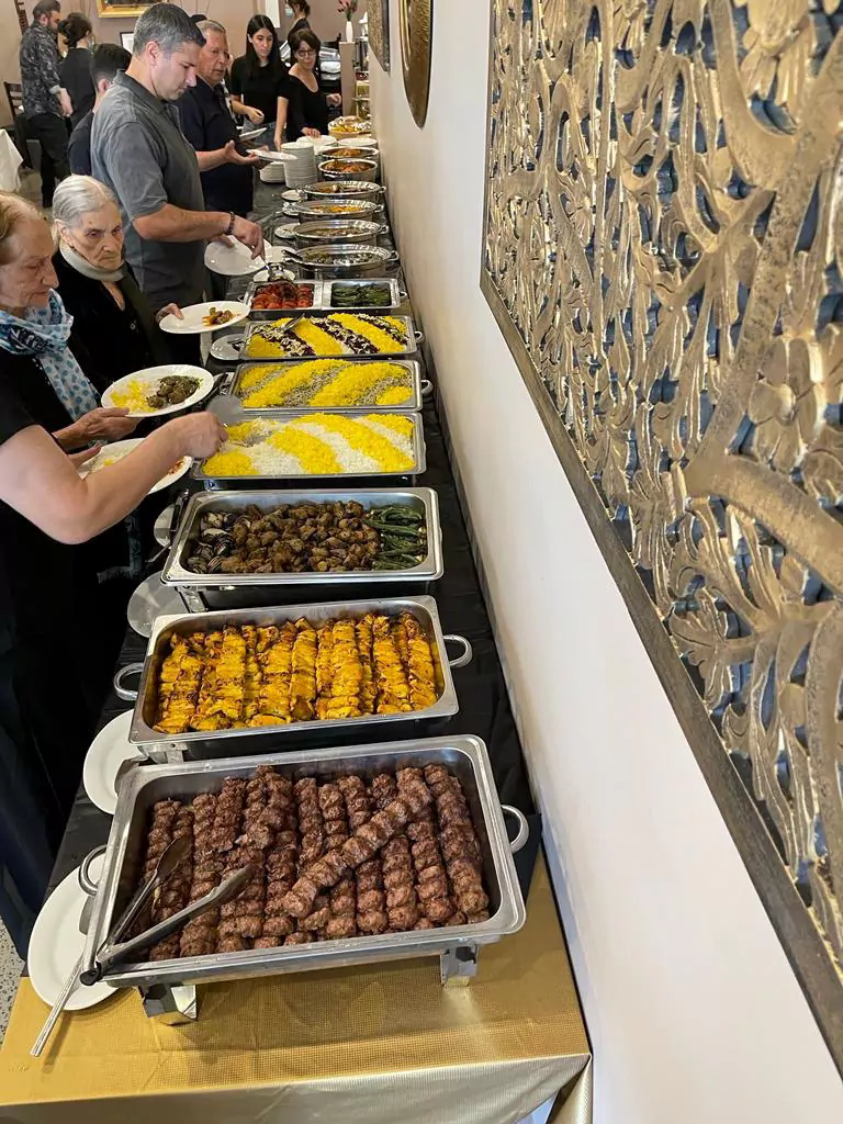 A line of guests serving themselves from an extensive Persian buffet catering at Narin Persian Restaurant in Sydney. The long table features stainless steel chafing dishes filled with authentic dishes, including grilled Koobideh and Joojeh kebabs, roasted vegetables, and beautifully decorated saffron rice with barberries, set alongside an ornate gold wall panel.