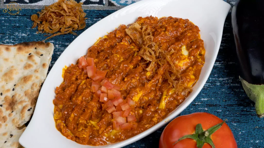Authentic Persian vegetarian Mirza Ghasemi made with smoked eggplants, tomatoes, and eggs, beautifully garnished with crispy fried onions and diced tomatoes, served in a white oval dish alongside flatbread at Narin Persian Restaurant.