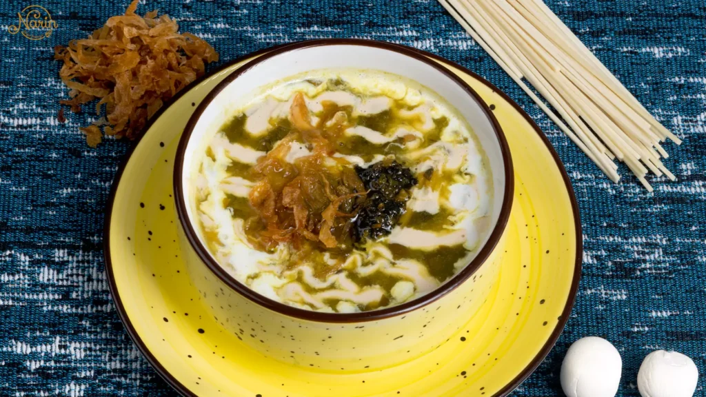 A hearty bowl of traditional Persian Ash Reshteh, a thick vegetarian herb and noodle soup, elegantly topped with liquid whey (Kashk), caramelized onions, and fried mint, served in a yellow bowl at Narin Persian Restaurant.