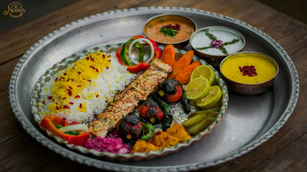 A Persian feast showing gas-grilled chicken kebab, a mound of Zereshk Polo with saffron and barberries, roasted vegetables, and side dips on a large silver platter