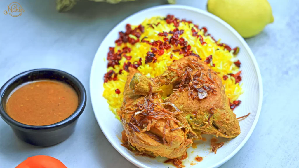 Close-up showing what is Zereshk Polo: a traditional Persian dish consisting of a chicken leg garnished with crispy onions, served alongside fluffy saffron rice, tart red barberries, and a small bowl of savory broth at Narin.   
